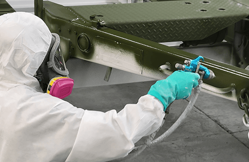 a spray technician in protective gear paints a part from a military vehicle