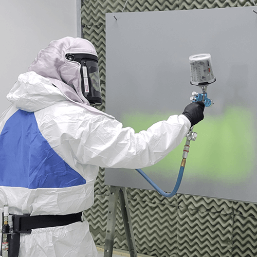 a paint technician in protective gear paints a metal panel inside a spray booth