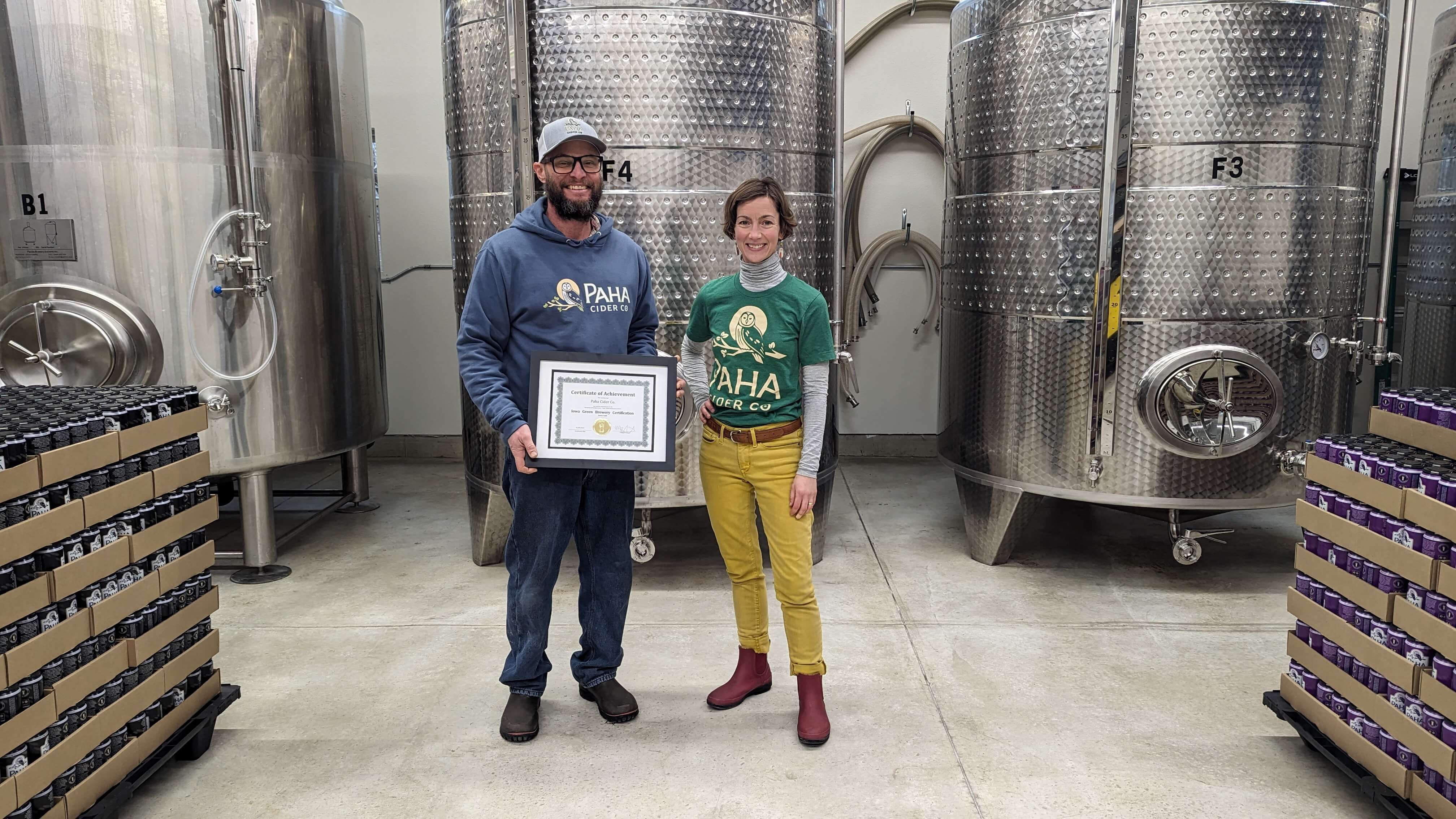 Paha Cidery Owner Mark Westbrock with Mo Volka, Sales & Marketing Director stand in front of cider vatts holding their Gold Certification