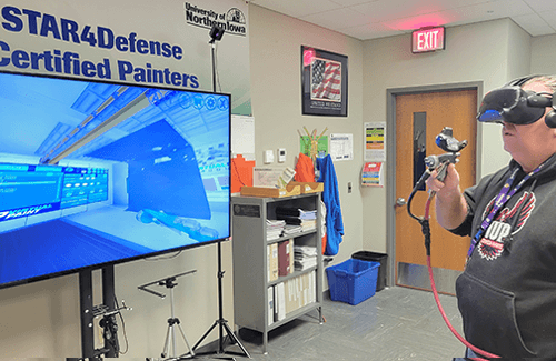 a man wearing a virtual reality mask and vr paint tool stands in front of a video screen showing a scene from VirtualPaint training simulation tool