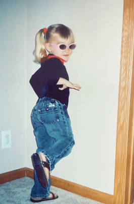 Young Kendall Lienemann, decked out in fashion forward jeans, black shirt, pink rimmed glasses, pigtails and red accents