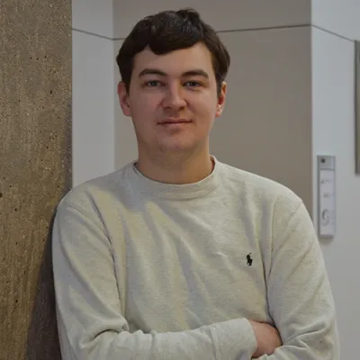 Young man in cream sweater leaning against a cement pillar